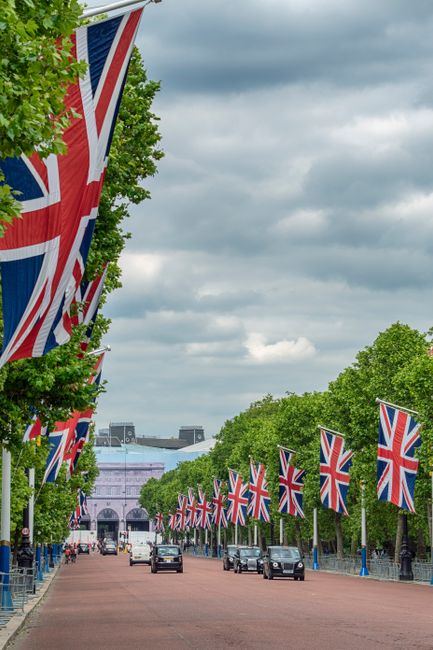 A wide street scene decorated with numerous Union Jack flags, which are attached along the route on tall black poles. The flags are prominently displayed, waving in the breeze against a backdrop of lu