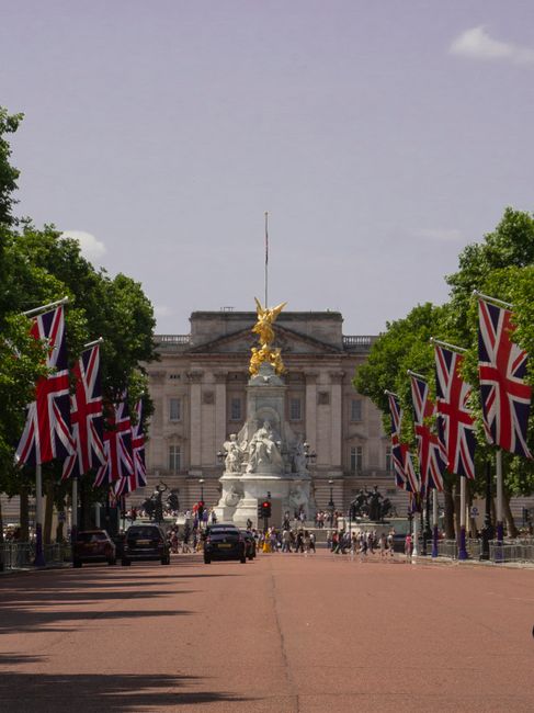 A wide street scene decorated with numerous Union Jack flags, which are attached along the route on tall black poles. The flags are prominently displayed, waving in the breeze against a backdrop of lu