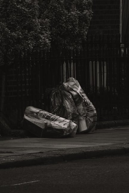 Two large black plastic rubbish bags are placed on the pavement next to a black iron fence, with one bag appearing more bulging and tangled than the other. The bags are filled with waste and are situa